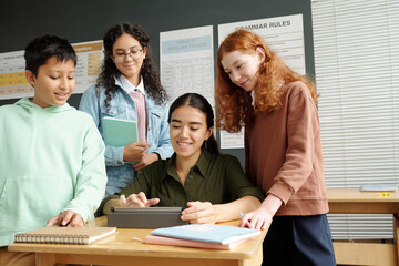 Fototapeta premium Young smiling teacher scrolling through online information on tablet screen while sitting by desk among clever learners during presentation