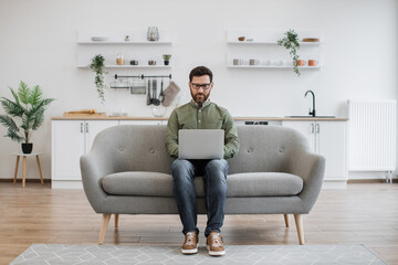 Obraz premium Focused caucasian man dressed in casual attire typing on wireless laptop while sitting on comfy sofa. Attractive male freelancer in eyeglasses doing online work while staying at home.