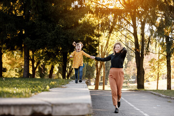 Mothers day. Happy young mom is holding by hand and walking with adorable little daughter at autumn park. Mom with kid are having fun, playing at weekend. Outdoors vacation activity. Children's day