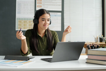 Confident young female teacher in headphones explaining grammar rule to online learner while sitting by workplace in front of laptop screen