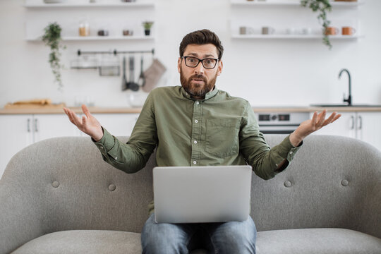 Confused bearded male wearing eyeglasses and casual attire shrugging shoulders while sitting on couch with modern laptop and looking at camera. Concept of doubt, people and technology.