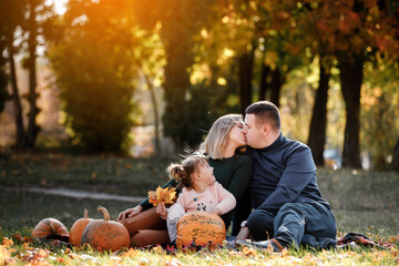 Family day. Happy mom and dad kissing little child daughter, having fun sit on grass with pumpkins at autumn park, enjoying spend time together at weekend. Parental care and happy carefree childhood