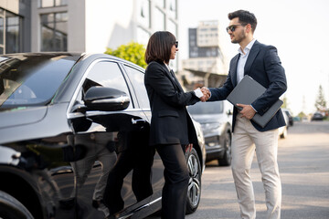 Business man and business woman shaking hands with each other, standing in front of the car outdoors. Concept of successful deal and partnership