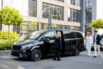 Female chauffeur waits a business people to let them in a minivan taxi, keeping door open. Concept of business trips and transportation service - 662948140