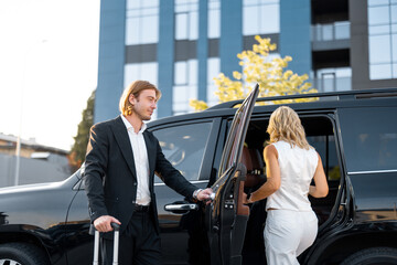 Elegant man helps a woman to get in the car, opening a vehicle door. Business couple having trip...