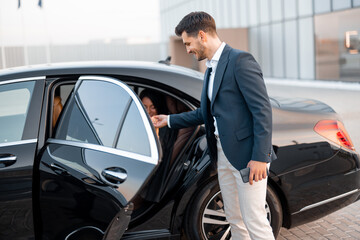 Businessman helps gently a woman to get out of a car, arrived by luxury vehicle for some event at evening. Concept of transportation and business lifestyle
