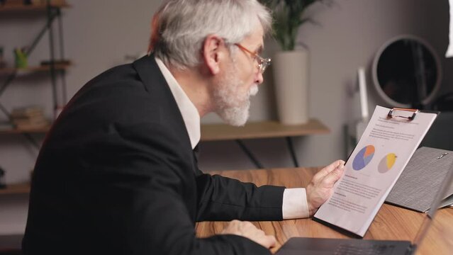 Side view of aged person wearing spectacles staring at balance sheet with charts before entering data on laptop. Thoughtful businessman in formal outfit analyzing sales data and market growth.