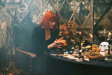 beautiful red haired young woman in black hat and witch outfit does magic in greenhouse decorated for Halloween celebration with pumpkins and skull in the smoke