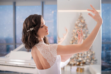 A ballerina in a white tutu poses in a bright hall with a Christmas tree.