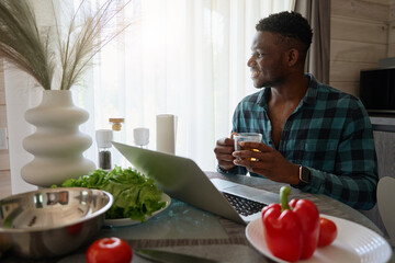 African American man sits in a cozy kitchen with laptop - obrazy, fototapety, plakaty