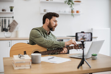 Concentrated guitarist sitting at table with wireless gadgets and holding guitar in hands while tuning instrument indoor. Attractive busy man shooting video tutorial on phone that standing on tripod.