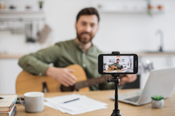 Fototapeta na wymiar Blurred background of positive male influencer doing live streaming while playing guitar at home office. Focus on tripod with modern smartphone and active screen.