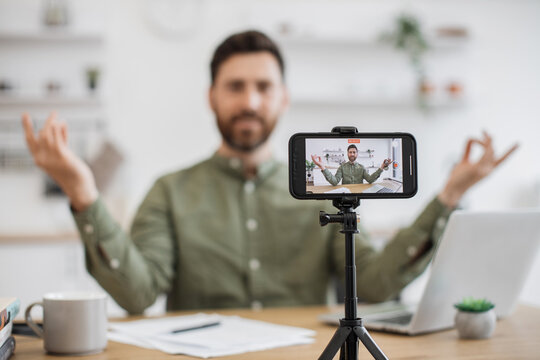 Popular cheerful blogger sitting at kitchen with wireless laptop and recording tutorial on back camera of mobile. Caucasian pleased man showing mudra gesture and vlogging meditation for subscribers.