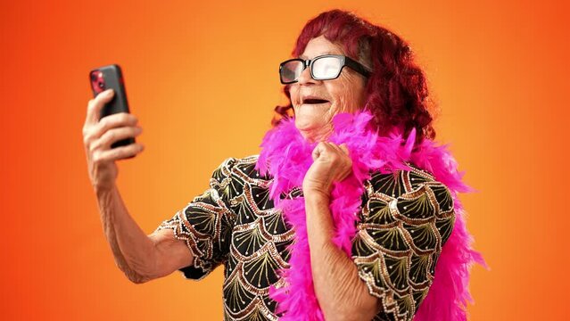 Funny crazy happy elderly old toothless woman taking selfie using mobile cell phone wearing boa and glasses isolated on solid orange background studio portrait. 