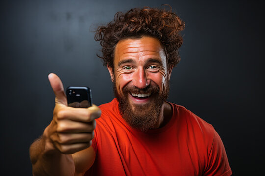 portrait of a expression of a  happy laughing man with brown hair and beard against colorful background who holds his thumbs up holding a smart phone
