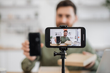 Focus on wireless smartphone standing on tripod and recording video. Cheerful male blogger showing and telling about modern gadget to subscribers in living room interior.