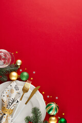 The art of elegance: preparing your Christmas table. Top view vertical photo of cutlery, plates, glassware, fir branches, сhristmas ornaments, stars confetti  on red background with promo spot