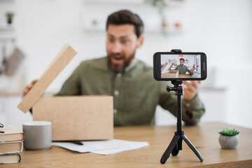 Caucasian bearded man making surprised facial expression while unpacking cardboard box during live streaming. Male influencer using modern smartphone and tripod for creating new content.
