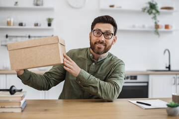 Excited caucasian man in casual wear and glasses sitting at home office with cardboard box in hands. Bearded male blogger happy to receive order from online store during holiday discount. - obrazy, fototapety, plakaty