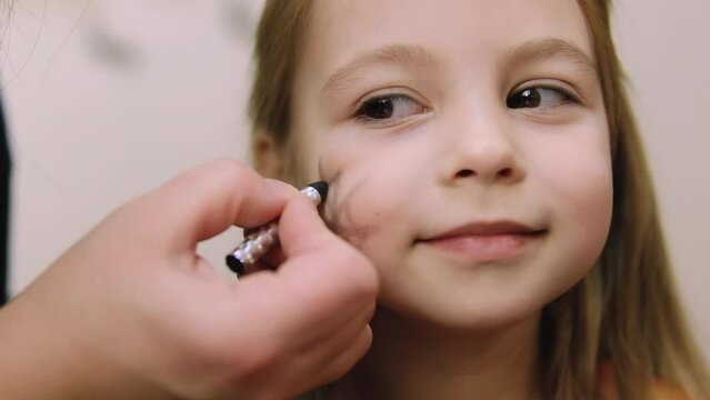 Unrecognizable woman doing eye makeup and drawing spider on face of Caucasian little girl sitting and waiting patiently