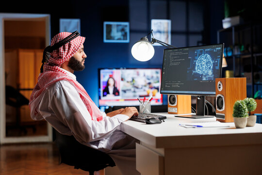 A professional man dressed in Arabic clothing, is working on futuristic interface while seated at his home office desk. Muslim guy manages data, monitors security, and programs AI on desktop computer.