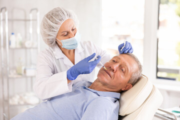 Obraz premium Experienced female cosmetologist working with senior male client in clinic of aesthetic medicine, performing minimally invasive procedure using filler injections to correct lips contour