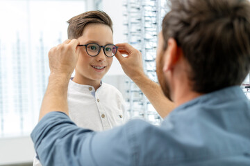 Authentic portrait of smiling son and father choosing and buying eyeglasses in modern optical store. Shopping, health care, eyesight concept