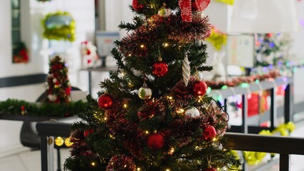 Close up shot of Christmas pine tree adorn with garlands and baubles in empty office, with blurry background of business charts on computer screens in xmas adorn workplace during winter holiday season