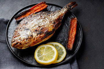 Close-up of baked dorado fish with lemon and carrots on black plate. Mediterranean cuisine