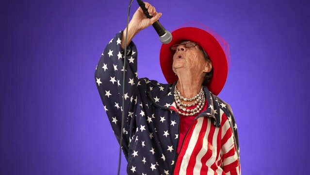 Funny crazy toothless grandmother with a fashionable look with glasses, singing enthusiastically into a microphone and dancing wearing US flag jacket isolated on solid purple background