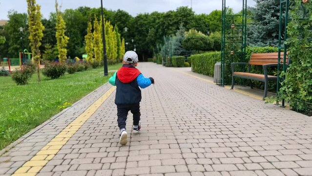 Happy kid in a jacket, cap and pants running by the park. Following the cheerful kid having active time outdoors.