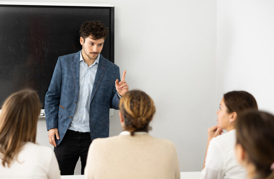 Young Male Teacher Near Interactive Board, Gesturing With Hand While Consecrating Topic Of Lesson For Adults In Classroom