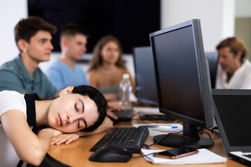 Young female developer tired after long workday napping near computer at office desk surrounded by coworkers..