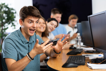 Excited young developer working at IT startup, watching project presentation with team gathered around table and using mobile phones for real-time data sharing