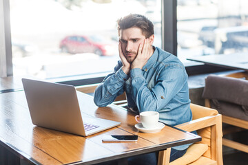 Portrait of upset stressed attractive young man freelancer in blue jeans shirt working on laptop, looking at display, expressing sadness and sorrow. Indoor shot near big window, cafe background.