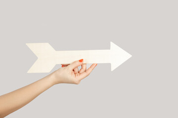 Closeup of woman hand holding white arrow pointing away, showing direction aside. Indoor studio shot isolated on gray background.