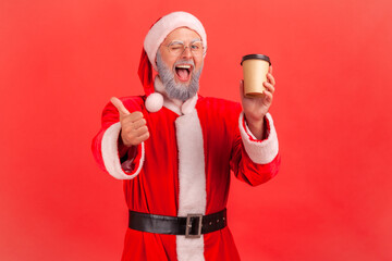 Fototapeta na wymiar Portrait of happy elderly man with gray beard wearing santa claus costume standing with disposable paper cup with coffee, showing thumb up. Indoor studio shot isolated on red background.