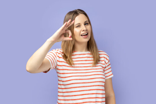 Portrait of cheerful young adult blond woman wearing striped T-shirt showing okay sign, assures you everything is fine, looks gladly. Indoor studio shot isolated on purple background.