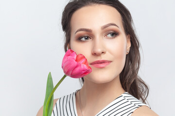 Obraz na płótnie Canvas Closeup portrait of romantic dreaming brunette woman standing looking away, holding pink tulip, enjoying aromat of flower, wearing striped dress. Indoor studio shot isolated on gray background.
