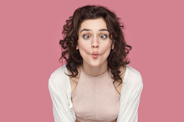 Fototapeta premium Portrait of funny positive woman with curly hair wearing casual style outfit having fun, making fish, demonstrates childish behavior. Indoor studio shot isolated on pink background.