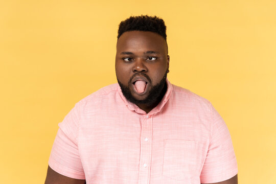 Portrait of disobedient crazy funny man wearing pink shirt showing tongue out and crossed his eyes displeased, behaving unruly naughty. Indoor studio shot isolated on yellow background.