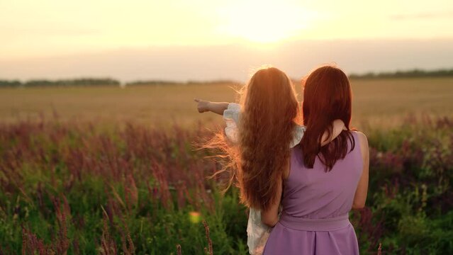 Little daughter points to horizon in mother arms at walk in field lit by sunset