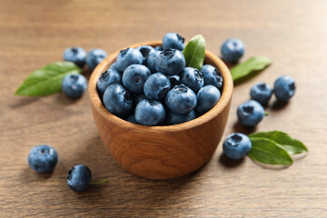 Bowl of tasty fresh blueberries with green leaves on wooden table, closeup
