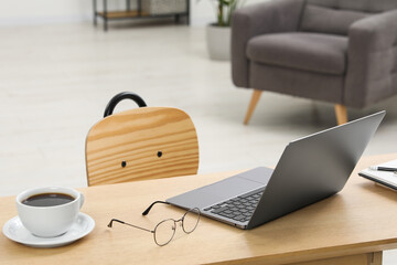 Home workspace. Laptop, glasses, cup of coffee and stationery on wooden desk in room. Space for text