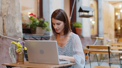 Pretty young woman working online distant job with laptop in city sunshine street cafe or restaurant browsing website chatting outdoors during break. Girl tourist looking at camera. Town lifestyles