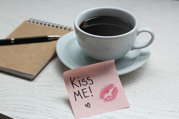 Sticky note with phrase Kiss Me, lipstick mark, cup of drink and notebook on white wooden table