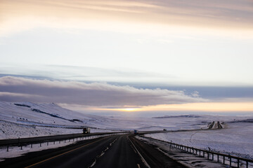 Beautiful winter landscape with a highway between blue snowy mountains, fluffy clouds on a sunset orange sky. Winter snow background. Elk Mountain, Wyoming, USA. Picturesque winter sky