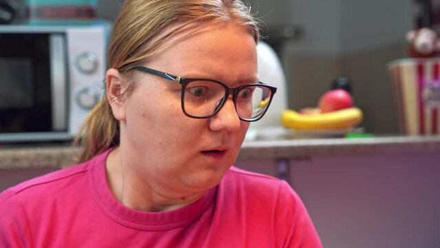 Young woman in glasses and pink T-shirt finds out shocking information while sitting in kitchen. Lady opens mouth in shock and slides down chair in gasp