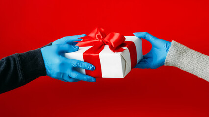 Pandemic gift. Present handing. Unrecognizable human hands in protective gloves holding wrapped box isolated on red background.