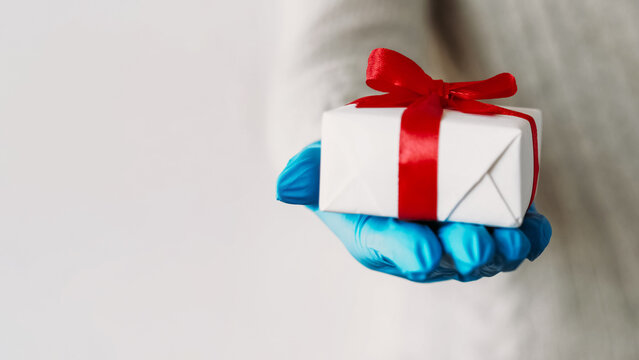 Pandemic gift. Virus present. Unrecognizable human hand in protective gloves holding wrapped box coronavirus protective gear isolated on gray copy space background.